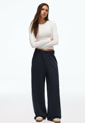 Mladá žena s dlhými hnedými vlasmi, ktorá má na sebe biely dlhý crop top s dlhým rukávom, voľné čierne nohavice a béžové topánky, stojí so zatvorenými rukami.
