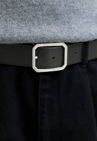 Schwarzer Ledergürtel mit einer achteckigen silbernen Schnalle, getragen über grauem Strickstoff und schwarzen Hosen. Strukturiertes Material und kontrastierende Farben sind sichtbar.