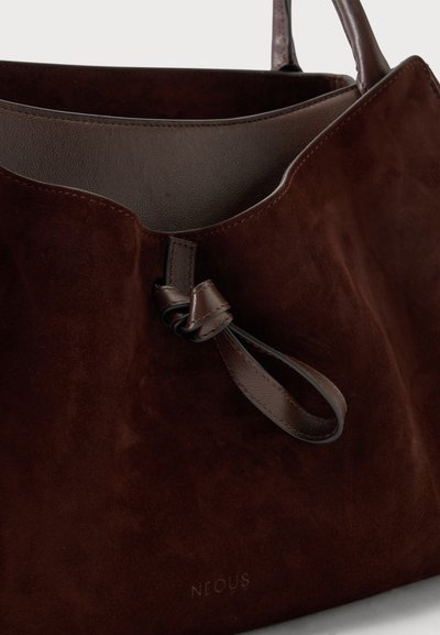 Sac fourre-tout en daim brun foncé avec poignée en cuir et détail de fermeture à boucle, montrant une doublure intérieure lisse et une gravure discrète du nom de la marque.