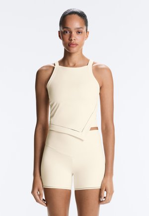 Jeune femme debout, portant un haut ajusté sans manches crème et un short taille haute assorti sur un fond clair uni.