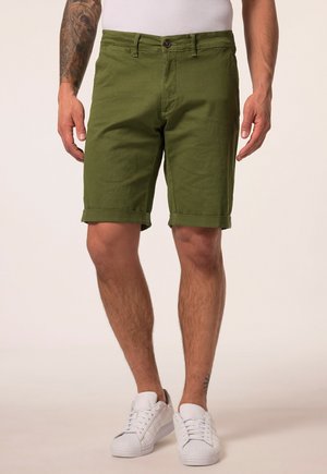 Mand iført grønne shorts med rullet kant, hvide sneakers og hvid ærmeløs top, stående med armene afslappet langs siden.