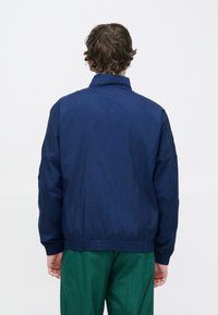 Temno modra vetrovka z visokim ovratnikom, elastičnimi manšetami in robom. Teksturirana tkanina z gladkim zaključkom, preprosta zasnova.