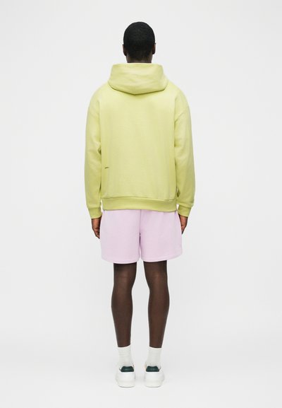 Άνδρας, εμφανιζόμενος από πίσω, φοράει ανοιχτό κίτρινο φούτερ με κουκούλα, ροζ παστέλ σορτς, άσπρες κάλτσες και άσπρα αθλητικά παπούτσια, με λευκό απλό φόντο.