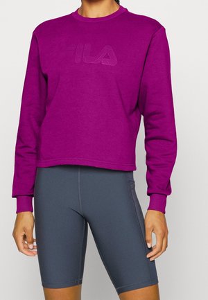 Persoon die een paarse FILA sweatshrit en grijze geribbelde fietsshorts draagt, staat met ontspannen armen tegen een effen achtergrond.