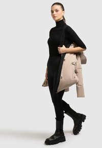 Maglione a collo alto nero, leggings neri e stivali alla caviglia spessi neri. Tiene in mano una giacca imbottita beige con una tracolla con logo.