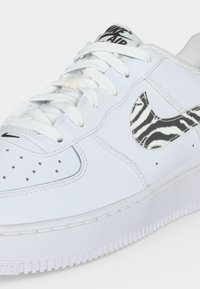 Λευκό αθλητικό παπούτσι Nike Air με λευκά κορδόνια και ασπρόμαυρο λογότυπο Nike swoosh με σχέδιο ζέβρας στο πλάι.