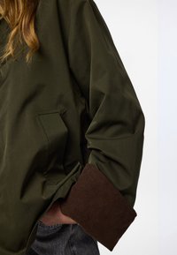 Olive roheline jakk, millel on pruun pits ja küljetasku. Kanga tekstuur on sile ja lõige on mugav.