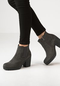 Stivaletti grigi alla caviglia con una finitura in suede testurizzata, pannelli laterali elasticizzati e un tacco spesso. Le suole nere presentano un design a tasselli per una maggiore aderenza.