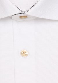 Gros plan d’un col de chemise blanche avec un bouton beige fixé et un second bouton beige cousu en dessous sur un tissu texturé.