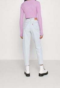 Kvinna i ljusblå högmidjade jeans, en kort lila ribbad tröja och vita snörstövlar, som står mot en enkel bakgrund.