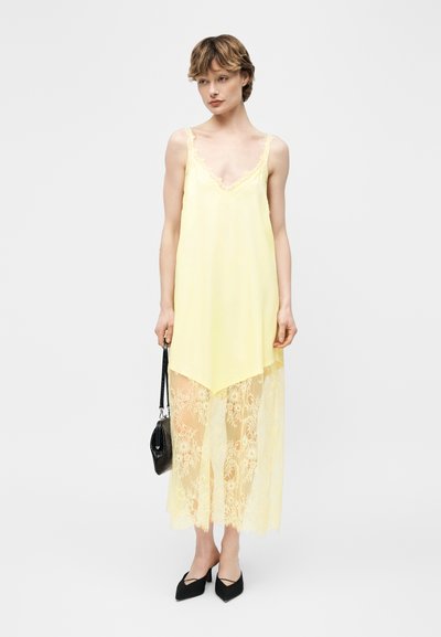 Νεαρή γυναίκα που φοράει ένα παλ κίτρινο φόρεμα-φορμάκι με δαντελένιο τελείωμα, κρατώντας μια μαύρη τσάντα χειρός και έχοντας μαύρες μυτερές γόβες, στέκεται σε λευκό φόντο.