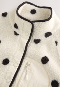 White fleece jacket with black polka dots, black trim on collar and front, snap buttons, and a zippered front pocket.