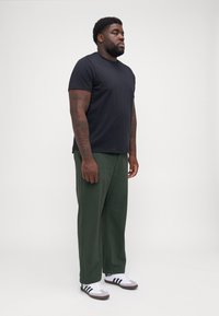 T-shirt noir à col rond associé à un pantalon vert ample et des baskets blanches avec des rayures noires. Le modèle se tient de profil contre un fond uni.