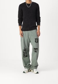 Personne portant un pull noir texturé, un pantalon vert large avec des motifs graphiques noirs, des baskets blanches et un collier chaîne en argent, debout immobile.