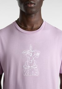 Camiseta de algodón de color lila claro con un gráfico blanco de un conejo sosteniendo un mando a distancia y la palabra "vans" debajo. Diseño clásico de cuello redondo.
