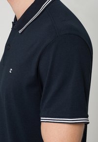 Homme portant un polo bleu marine avec un col et des manches à rayures blanches, et un petit logo brodé sur la poitrine.