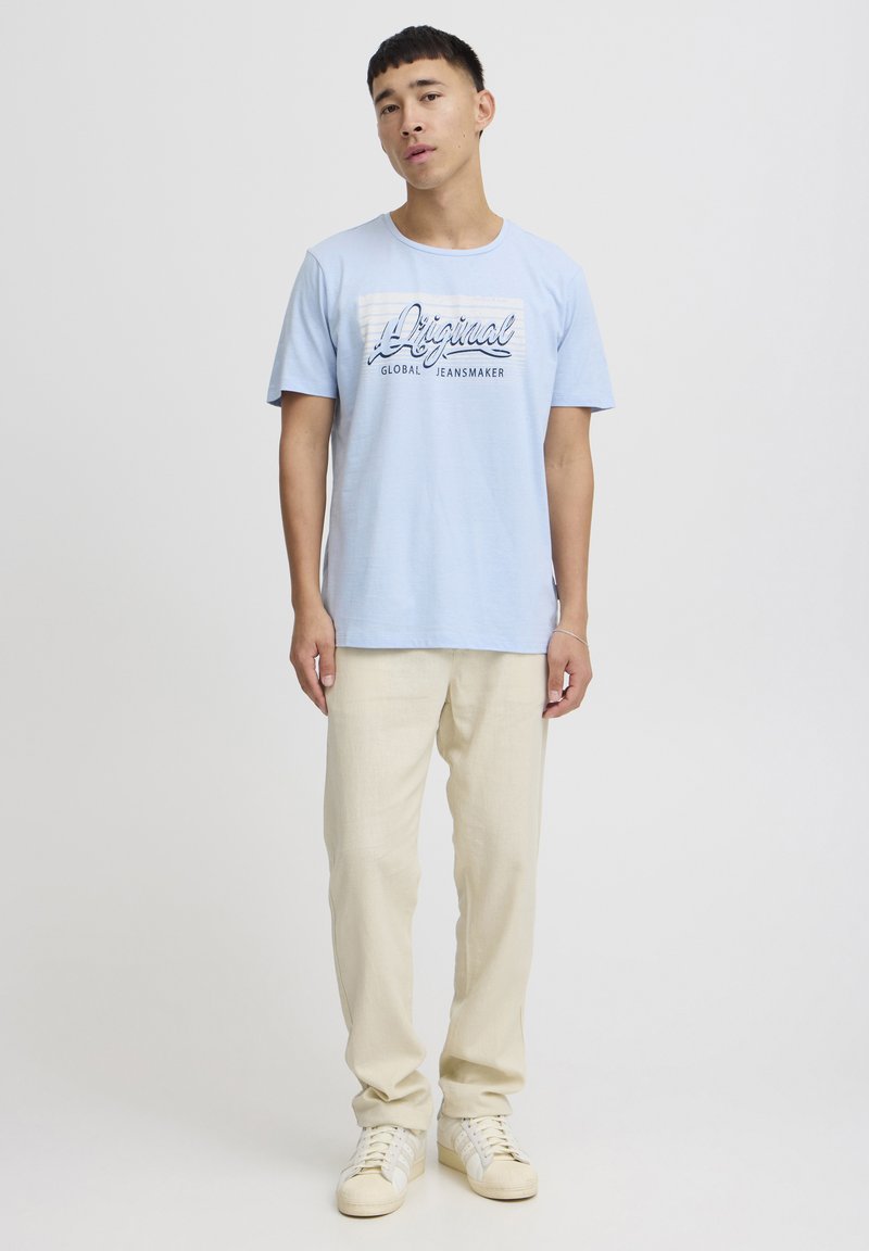Lys blå bomull T-skjorte med "Original Global Jeansmaker" grafikk, kortermet, sammen med lyse beige bukser og hvite sneakers.