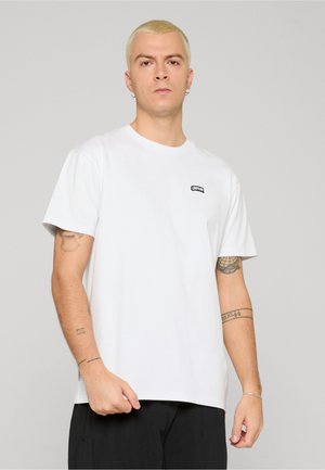 Joven con cabello corto rubio, vistiendo una camiseta blanca con un pequeño logo negro que dice "UNFAIR", pantalones negros y tatuajes visibles en el brazo.