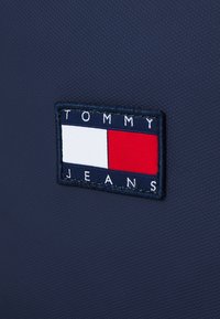 Étiquette en tissu Tommy Jeans avec un logo rectangulaire bleu marine, blanc et rouge sur un fond textile texturé foncé.