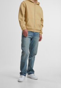 Beiger Kapuzenpullover mit Reißverschluss und Kängurutasche, kombiniert mit hellblauen Jeans und weißen Turnschuhen. Schlichtes Design, keine sichtbaren Muster oder Verzierungen.