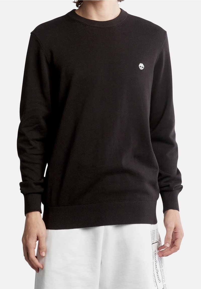 Personne portant un pull noir à col ras du cou à manches longues avec un petit logo d'arbre blanc sur le côté gauche de la poitrine et un short blanc avec un texte noir.