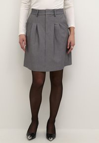 Jupe plissée grise avec une taille structurée, texture du tissu lisse, complétée par des collants noirs transparents et des talons noirs brillants avec des accents métalliques.