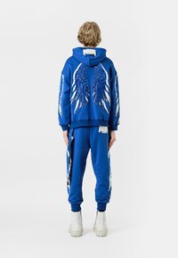 La sudadera azul y los pantalones deportivos presentan diseños de alas blancas en la parte posterior y los laterales. La ropa está hecha de un material suave con puños de canalé.