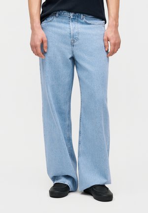 Άνδρας που φοράει ανοιχτό μπλε τζιν με φαρδιά μπατζάκια και μαύρα παπούτσια χωρίς κορδόνια, στέκεται μπροστά σε ένα απλό λευκό φόντο.