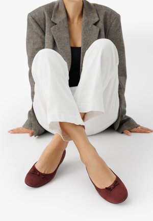 Donna seduta sul pavimento con pantaloni bianchi, blazer grigio, top nero e ballerine borgogna con piccoli fiocchi.