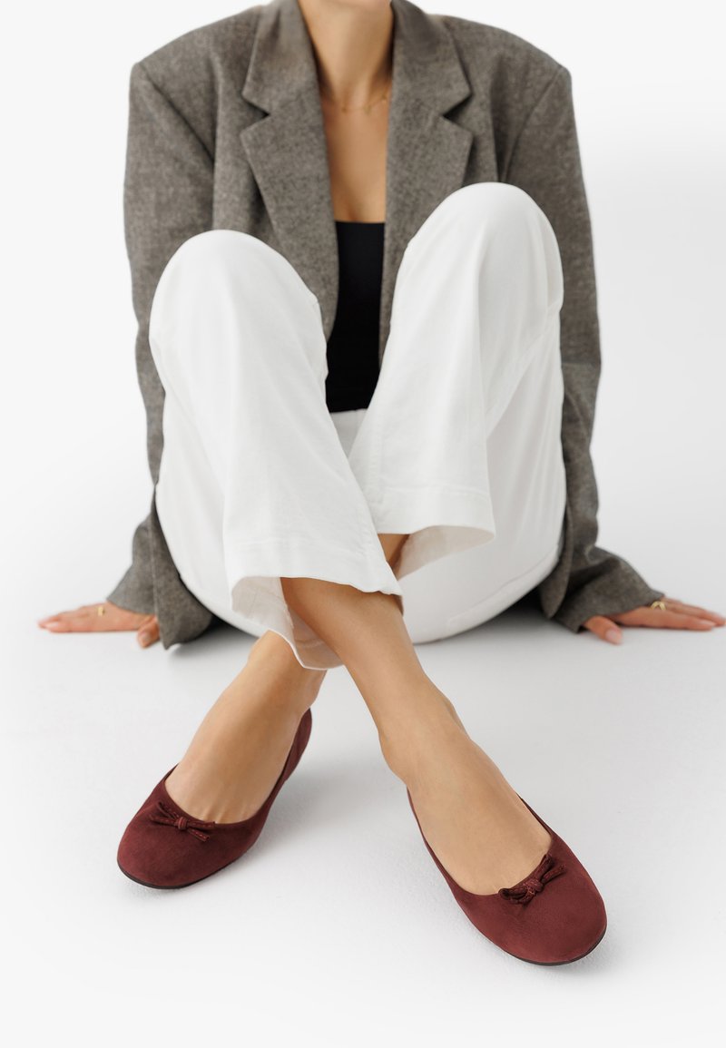 Sieviete, kas sēž uz grīdas, ģērbta baltos biksēs, pelēkā žaketē, melnā blūzē un bordo krāsas balerīnās ar maziem ziediem.