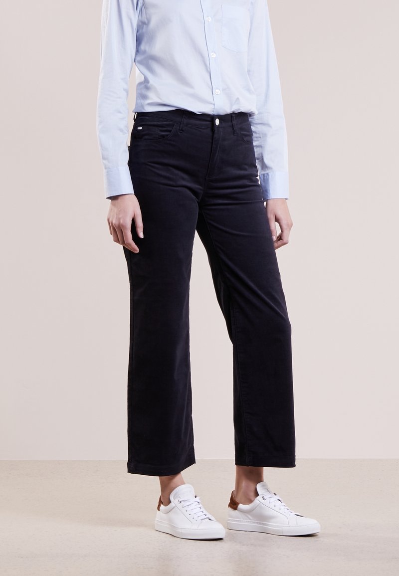 Personne portant un pantalon noir droit, une chemise blanche boutonnée et des baskets blanches, debout devant un fond uni.