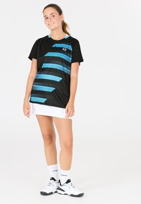 Svart träningssko med blå randig design, kombinerad med en vit kjol. Dekorativ logotyp på tröjan. Modell med vita skor och strumpor.