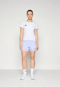 Wit sportief t-shirt met zwart logo, gecombineerd met lichtpaarse shorts. Draagt witte sokken en schoenen, en straalt een sportieve uitstraling uit.