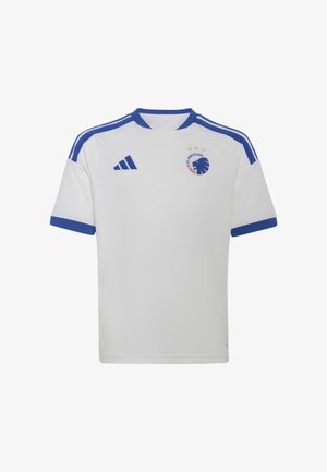 Un jeune garçon se tient avec assurance, tenant un ballon de football, portant un équipement de football blanc et bleu avec des chaussettes et des crampons assortis sur un fond uni.