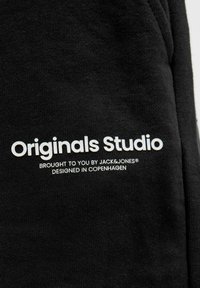 Crni pamučni materijal s glatkom teksturom, s bijelim otisnutim tekstom: "Originals Studio," "Dovela vas marka JACK & JONES," i "Dizajnirano u Kopenhagenu."