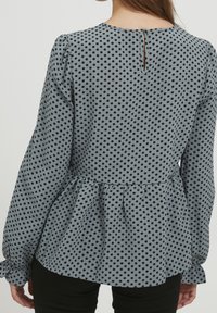 Blusa grigia con pois neri, vita peplum, maniche lunghe e dettaglio a chiave sulla schiena. Tessuto morbido con leggero arricciamento ai polsini delle maniche.