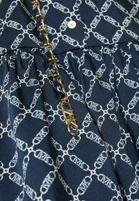 Tissu bleu marine avec motifs géométriques de chaînes blanches, bouton doré et bandoulière chaîne dorée avec maillons entrelacés et détail de fermoir triangulaire.