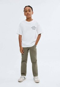 Ett barn står med en hand i fickan iförda en vit "Notes Music Fest" T-shirt, olivfärgade byxor och vita sneakers framför en neutral bakgrund.