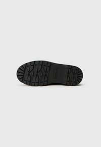 Suela de zapato de goma negra con patrón de textura, que presenta una punta redondeada y un pequeño detalle de marca cerca del talón.