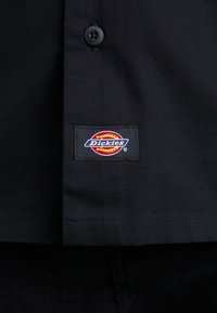 Vêtement noir avec un bouton et un écusson rectangulaire du logo de la marque Dickies en rouge, jaune et blanc cousu près de l'ourlet.