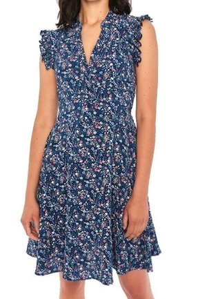 Frau trägt ein ärmelloses blaues Blumenkleid mit Rüschen an den Schultern und einem V-Ausschnitt, steht mit entspannten Armen seitlich am Körper.