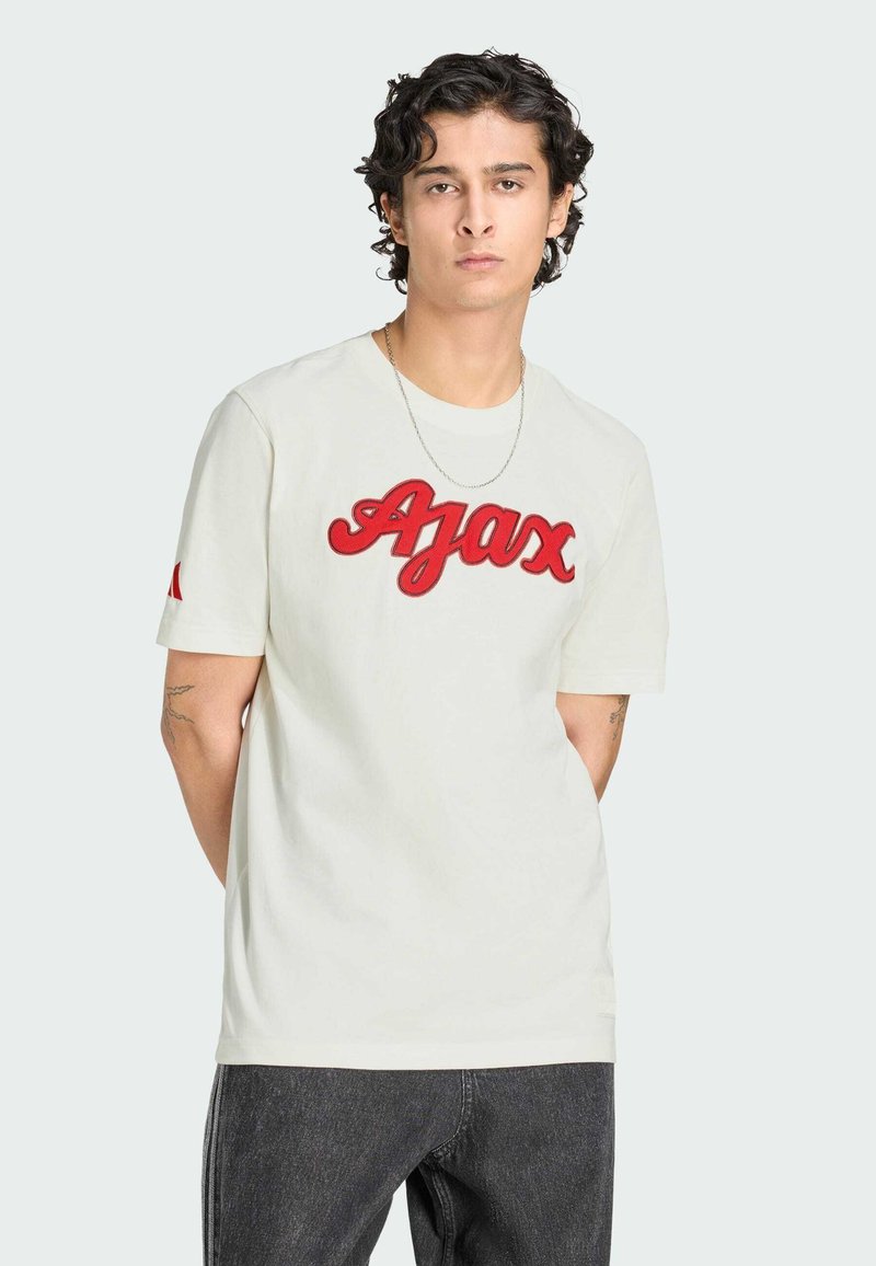 Jeune homme aux cheveux bouclés et foncés portant un t-shirt blanc avec le texte rouge "Ajax" et un jean foncé, debout les mains derrière le dos.