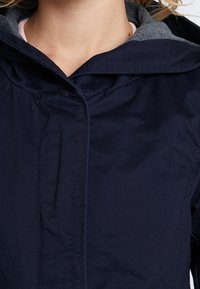 Marineblauwe jasstof met een grijze voering aan de binnenkant. Voorzien van een capuchon, knoopsluiting en een gladde textuur met minimale zichtbare details.