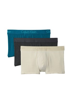 Gespiereerde man staat op blote voeten met handen achter rug, gekleed in turquoise Calvin Klein boxershorts tegen een effen grijze achtergrond.