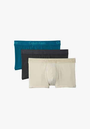 Homme musclé debout pieds nus, mains dans le dos, portant un boxer bleu sarcelle Calvin Klein sur un fond gris uni.