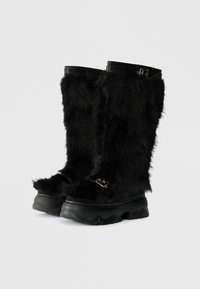 Črne škornje iz krzna z debelim, teksturiranim dizajnom, ki vključuje sijoče črne detajle in masiven gumijast podplat. Podrobnosti s hardwareom vključujejo sponke.