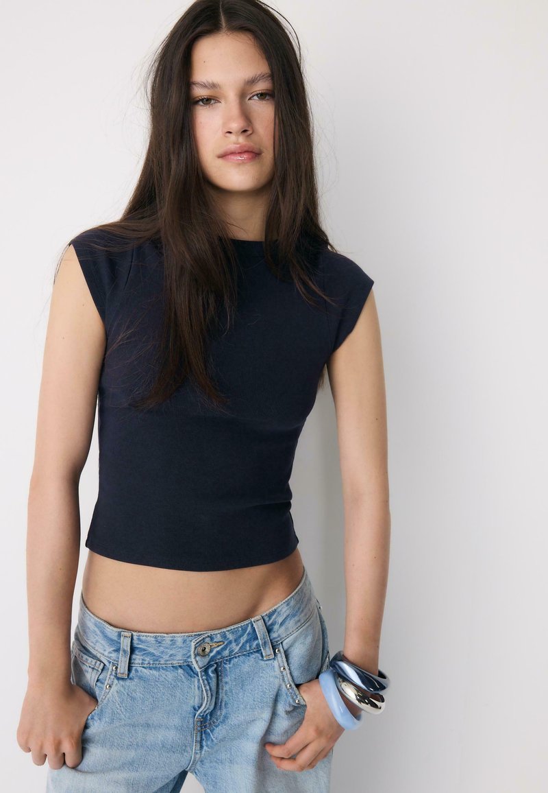 Jeune femme aux longs cheveux bruns portant un haut court noir sans manches, un jean bleu clair et des bracelets bleus et argentés à son poignet droit.