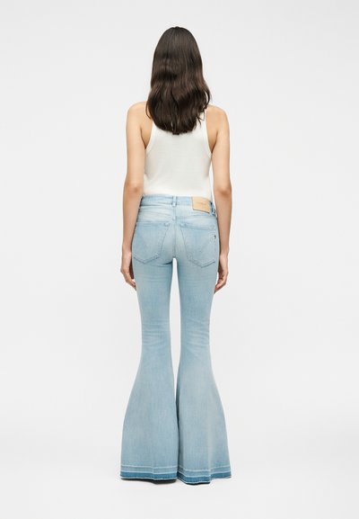 Γυναίκα με μακριά σκούρα μαλλιά που φοράει άσπρη φανέλα και ανοιχτό μπλε τζιν ψηλόμεσα και με φαρδιά μπατζάκια, στέκεται γυρισμένη με την πλάτη μπροστά σε λευκό φόντο.