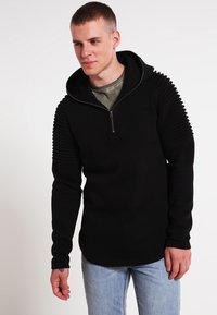 Svart zip-up hettegenser med ribbede detaljer på skuldrene og ermene, kombinert med en lys grønn skjorte under og blå jeans. Smooth tekstur.