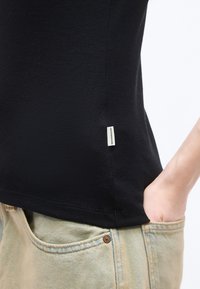 Κοντινό πλάνο ενός ατόμου που φοράει μαύρο πουκάμισο με μια μικρή λευκή ετικέτα και ανοιχτόχρωμα τζιν τζιν με το ένα χέρι στην τσέπη.